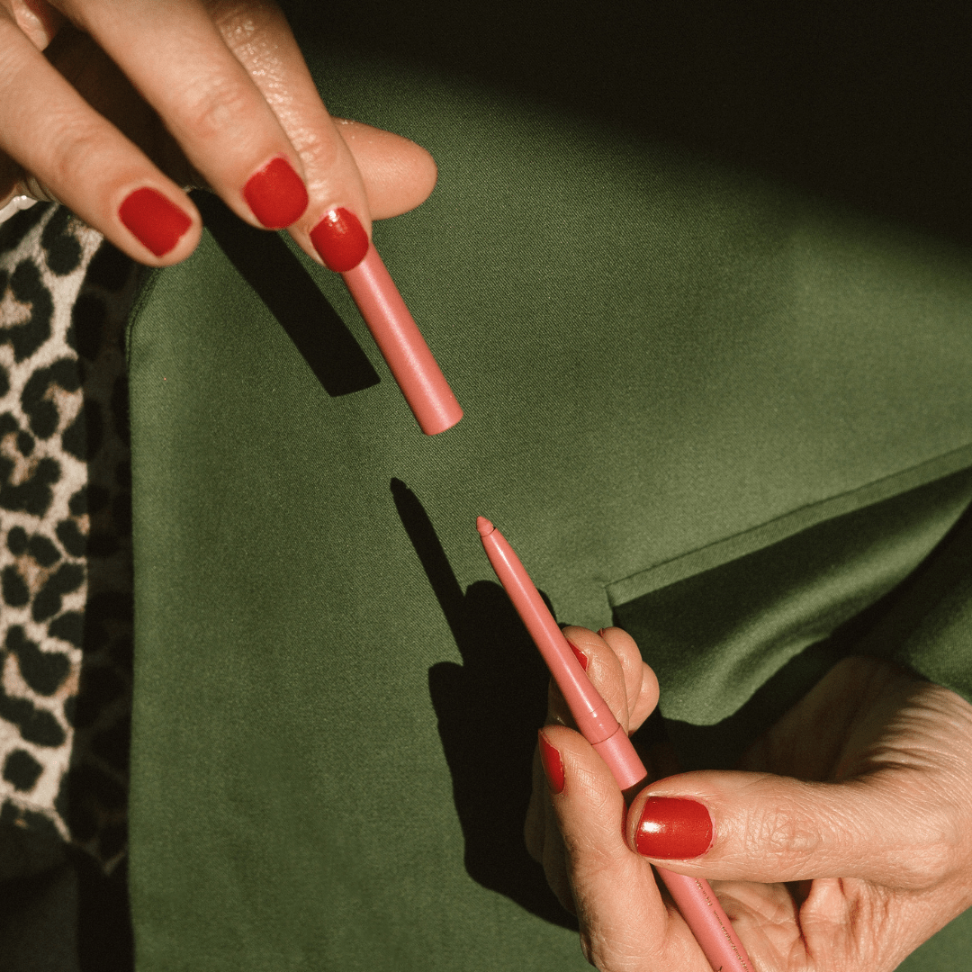 Close-up of hands with red nail polish holding Eco by Sonya nude lip liner against a green background